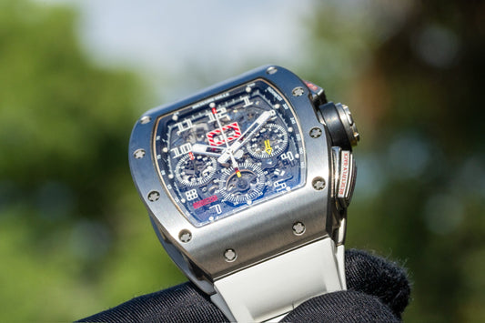 A close-up of a Richard Mille chronograph watch with a skeleton dial and white strap, held by a gloved hand against a blurred green outdoor background.