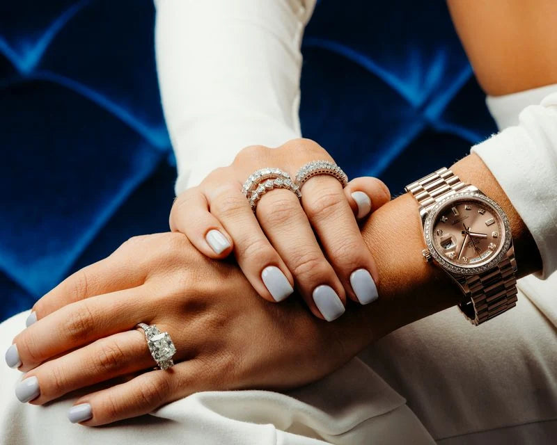A model wearing diamond rings and a rose-toned watch against blue upholstery.