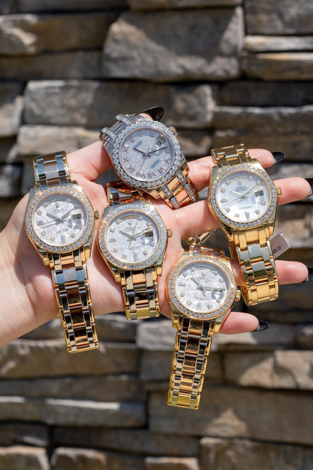 A hand holding five luxury Rolex watches with gold and diamond details, showcasing elegant craftsmanship and premium design against a stone wall background.