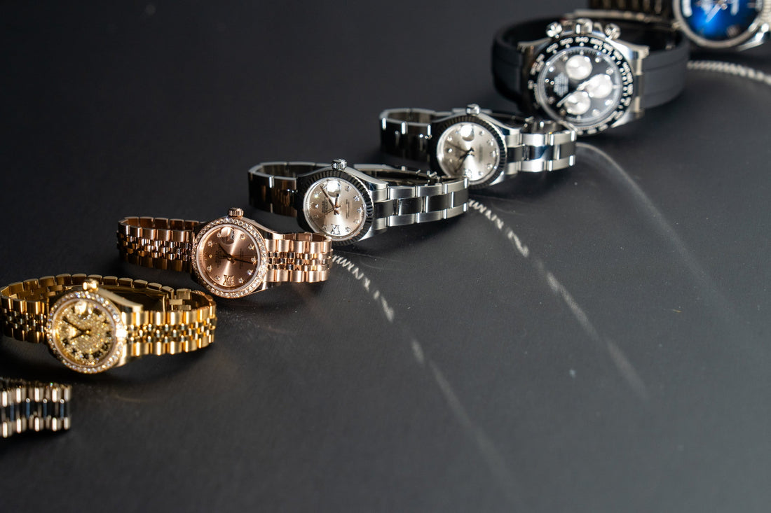 A collection of luxury wristwatches displayed in a diagonal arrangement on a dark surface, featuring gold, rose gold, and stainless steel designs with diamond accents and polished bracelets.