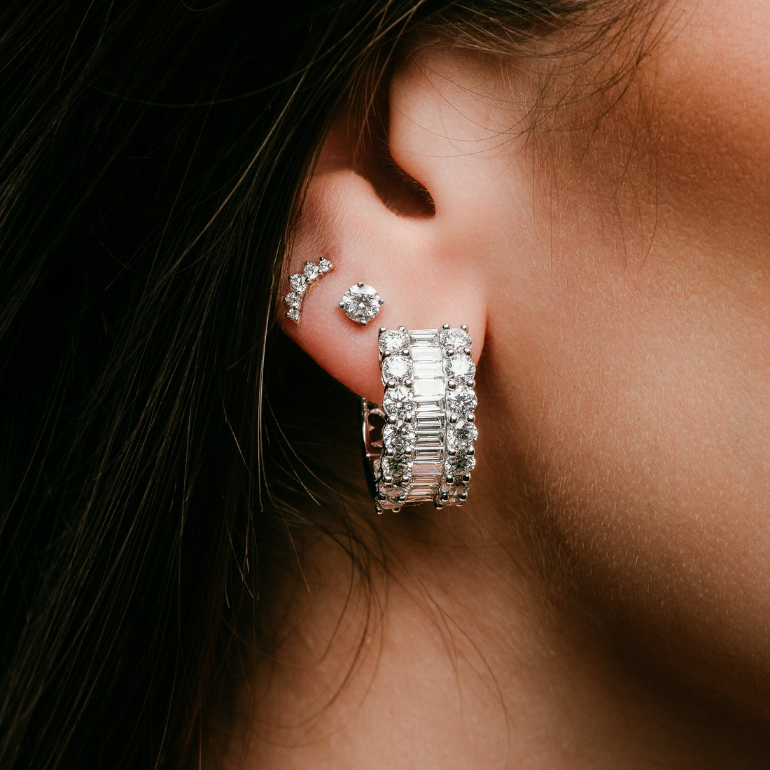 A close-up of an ear featuring a curated jewelry stack, including a wide diamond hoop with baguette and round stones, a classic diamond stud, and a delicate curved diamond crawler.