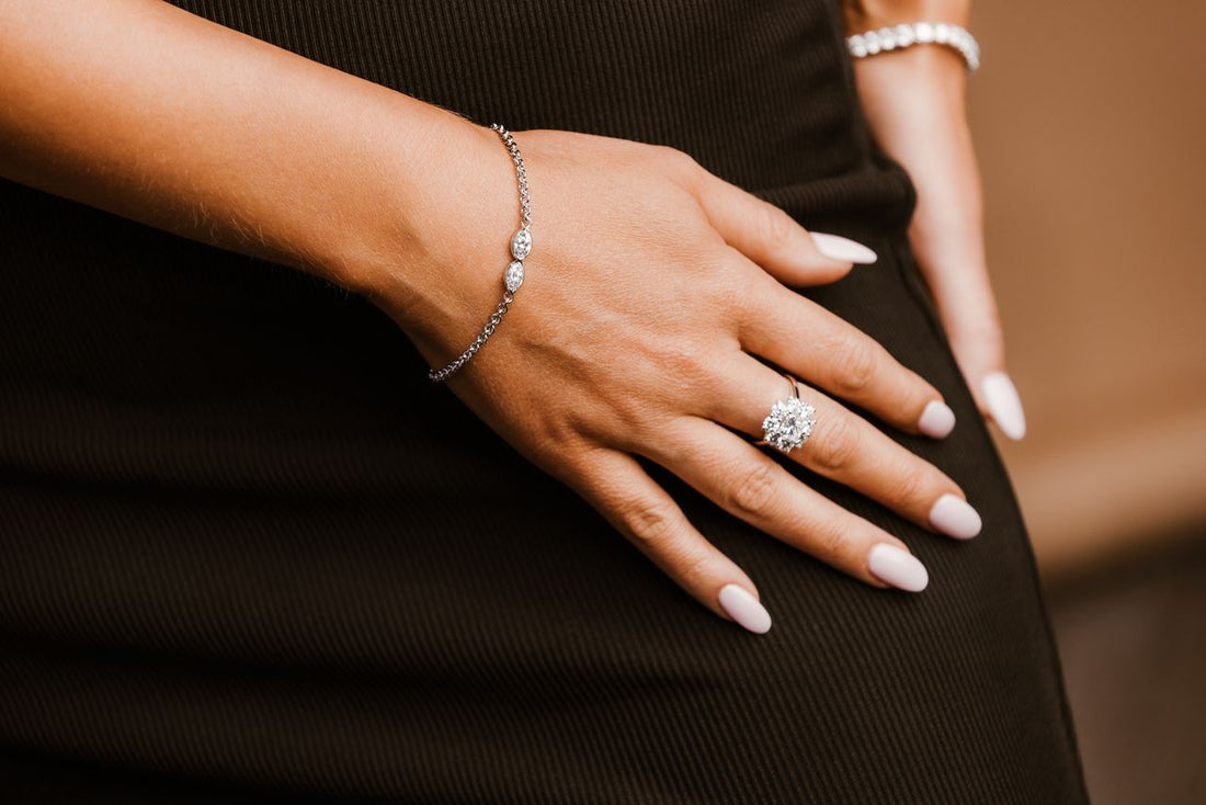 A person's hand resting on a dark garment, featuring a silver chain bracelet with three oval stones and a large cluster diamond ring.