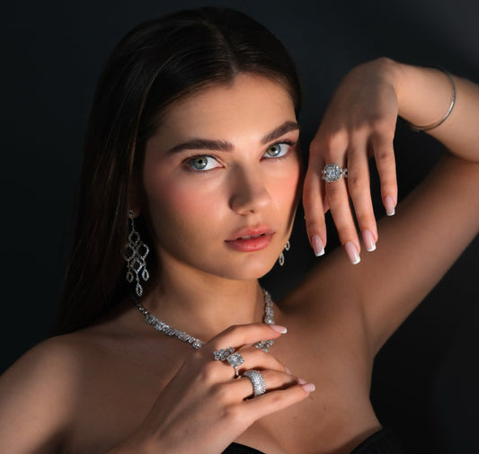  A striking close-up photograph of a woman wearing an extensive set of diamond jewelry, including chandelier earrings, a statement necklace, and multiple rings, posing dramatically against a dark background.