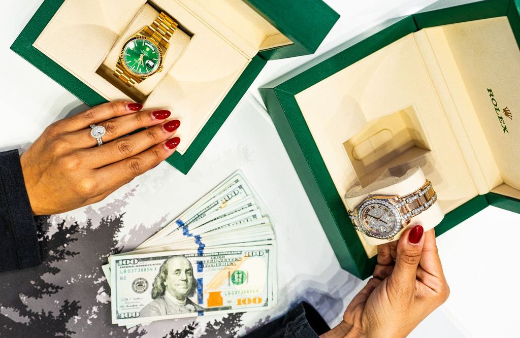 Hands with red nails display two Rolex watches in green boxes—one gold with a green dial and one diamond-encrusted—alongside a stack of hundred-dollar bills.