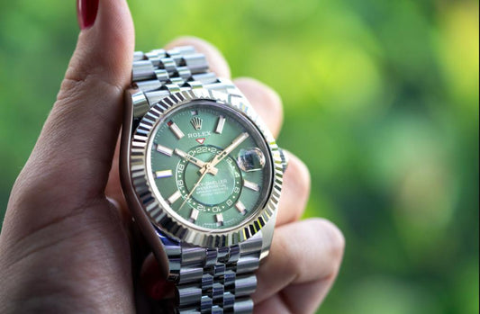 Close-up of a person holding a Rolex Sky-Dweller watch with a green dial and stainless steel bracelet, featuring a fluted bezel and date display under natural light.