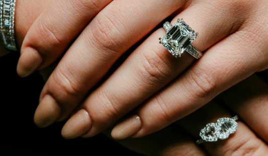 An emerald cut center stone ring worn beside a ring with two halo settings.