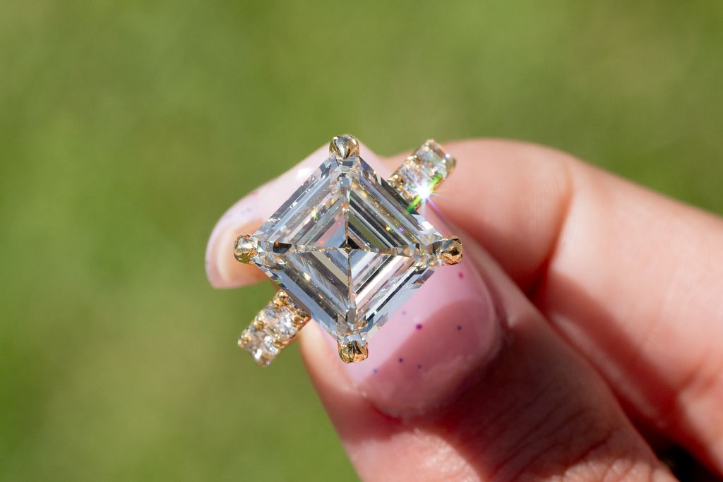 Asscher-cut or princess-cut diamond ring with a yellow gold band and diamond accents, held between two fingers.