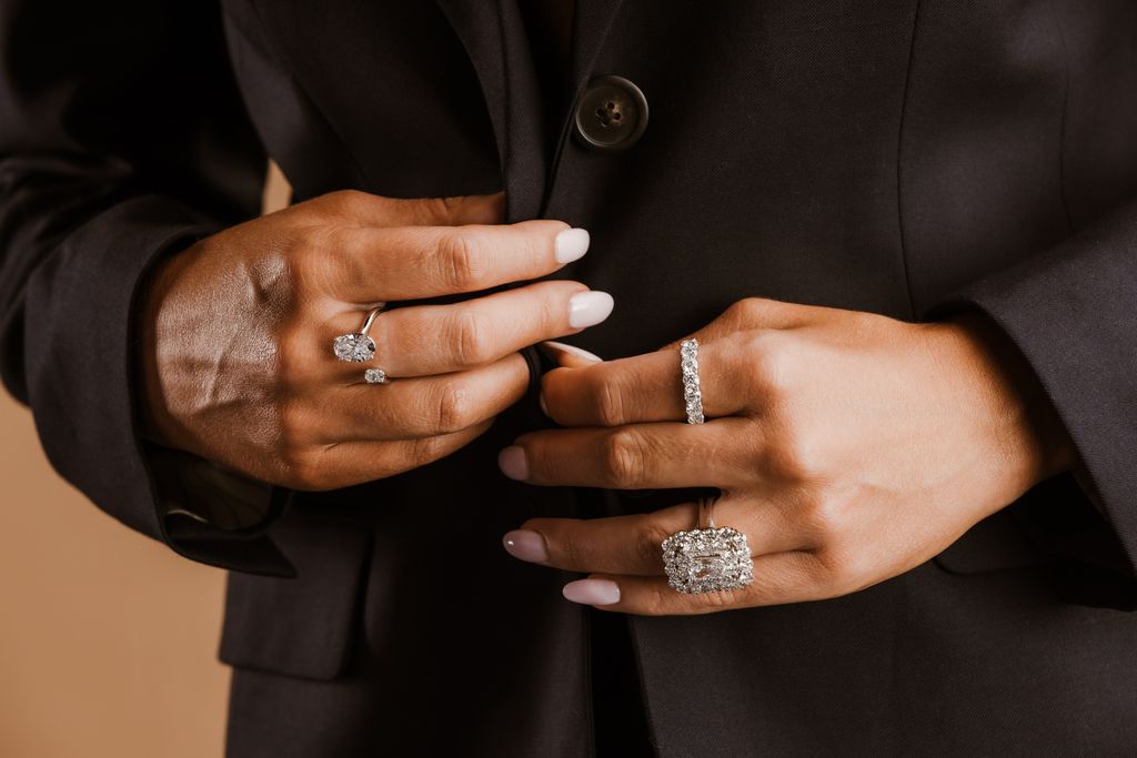 A person wearing a black blazer displays multiple diamond rings, including a unique open-band toi et moi ring and a large emerald-cut halo ring.