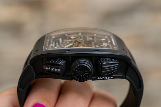 Close-up side profile of a luxury black watch, the Richard Mille RM 011-FM, showing the crown, pushers, and black rubber strap.