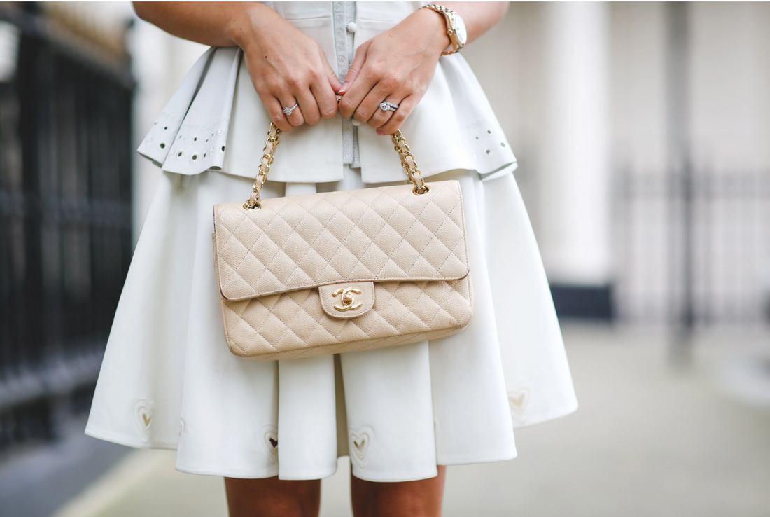 A mid-shot of a person wearing a white dress, holding a quilted beige Chanel Classic Flap bag with a gold chain strap, also showing a rose-gold-toned watch and rings on their fingers.