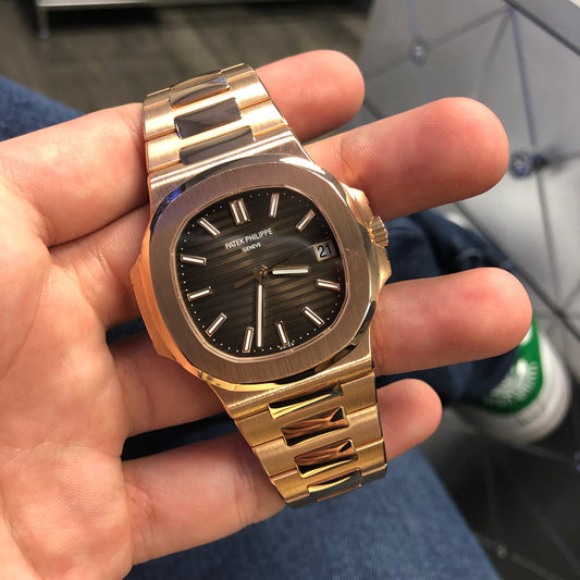 A gold tone Patek Philippe Nautilus with a dark dial, held in one hand.