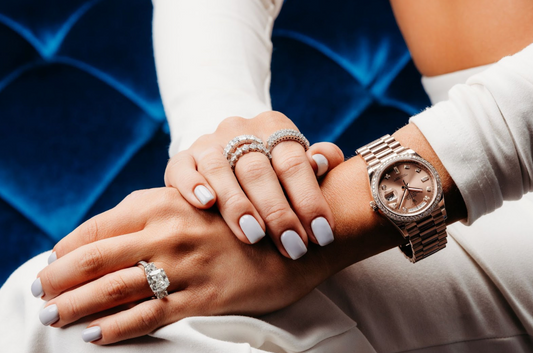 Elegant woman wearing a diamond Rolex watch and multiple diamond rings with white manicure, resting hands on blue velvet background.