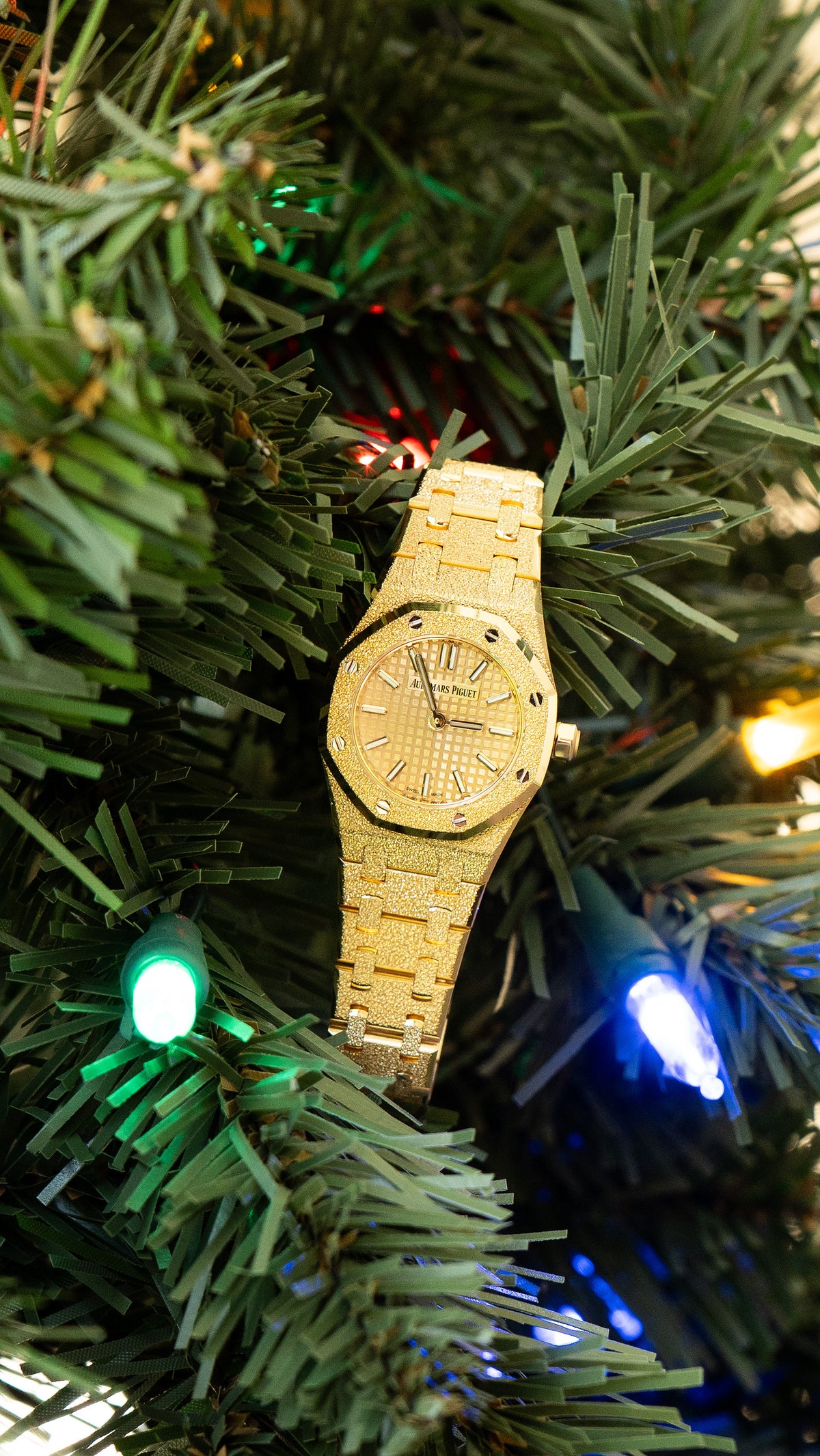 Gold tone Audemars Piguet watch with a textured dial and bracelet among green branches and lights