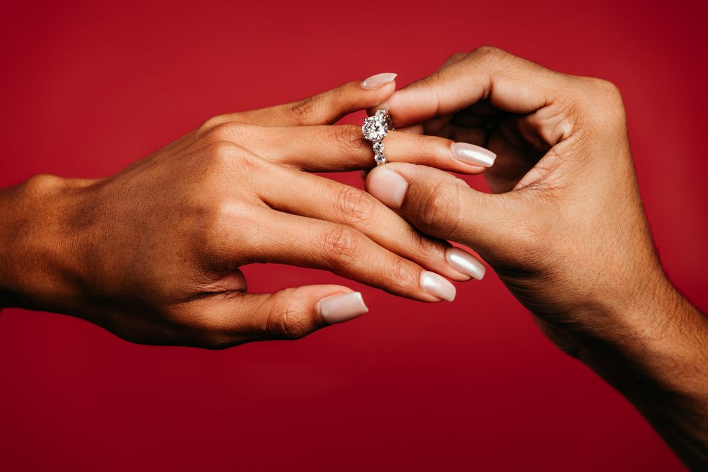A ring with a large center stone being placed on a finger against a red background