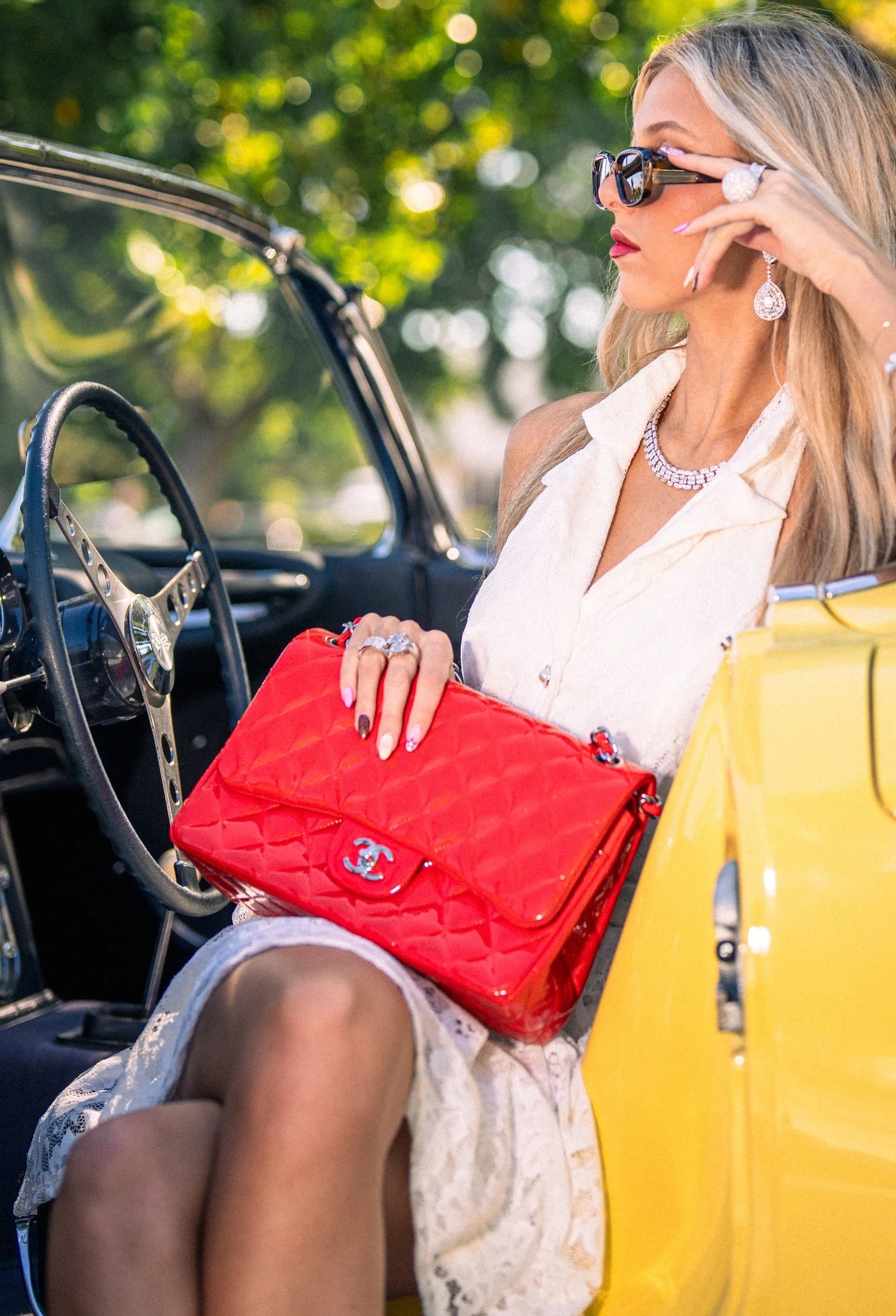 Model wearing jewelry and holding a red handbag in a yellow convertible