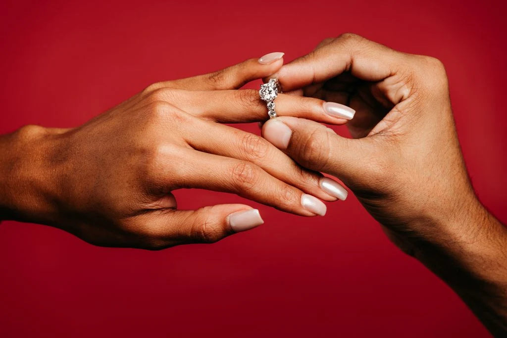 A ring with a large center stone being placed on a finger against a red background