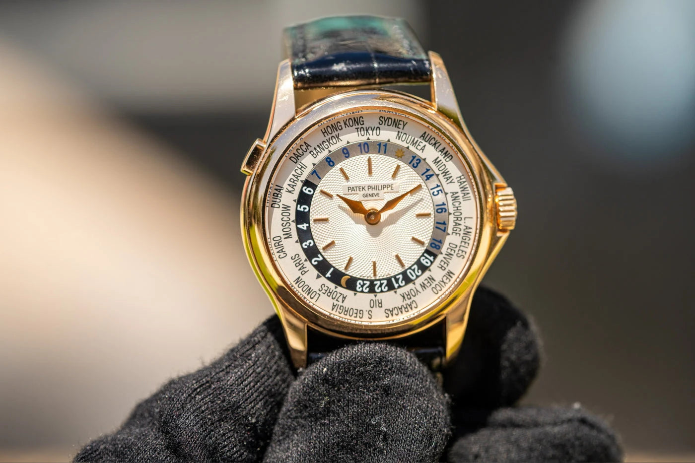A gloved hand holding a gold Patek Philippe world-time watch with a leather strap.